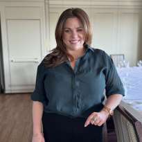Woman, Shoulder length hair, blueish shirt black pants standing next to a chair