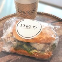 A chicken salad sandwith with greens, wrapped in cellophane and sealed with a Davio's sticker, accompanied by a to go cup of coffee with a Davio's cardboard sleeve