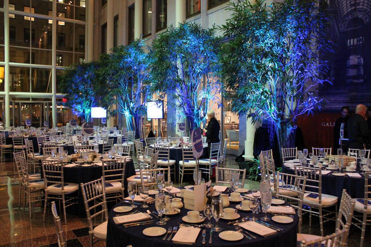 High ceilinged lobby space set with large round tables and blue light illuminating trees in planters