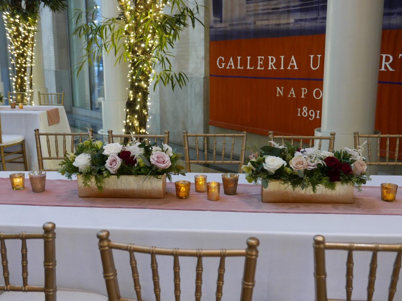 Closeup of a long table with rose centerpieces, gold chairs, and votive candles. Trees wrapped with fairy lights in the background.