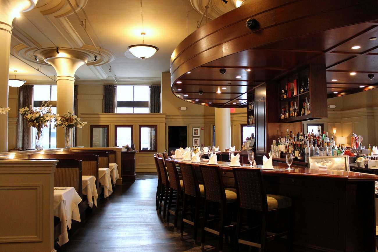 View of the main dining room with booths on the left and oval-shaped bar on the right. The room features high ceilings with pendant lights, tall windows, and Davio's signature columns. 