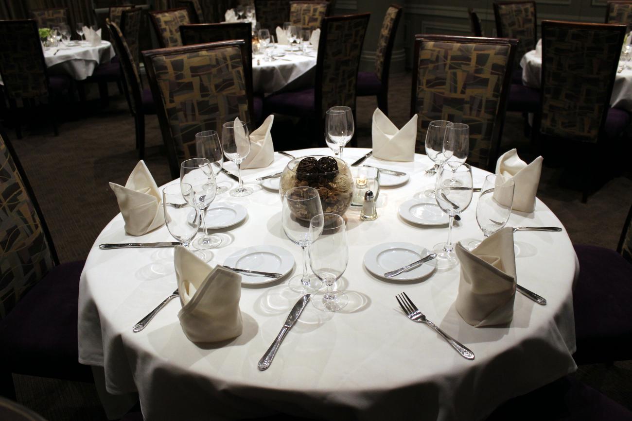 A round table set for 6, with white tablecloth, cloth napkins, silverware, stemware, and bread plates. Similar round tables are seen in the background.