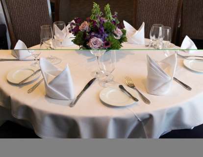 Closeup of a round table set for six, with white tablecloth, cloth napkins, and wineglasses. The centerpiece is an arrangement of lavender roses, purple foxgloves, purple hydrangea, and greenery.