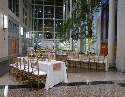 Long tables with white tablecloths and gold chairs, white floral arrangements, potted trees wrapped with white lights, and a stories-high window create the perfect setting for a holiday event