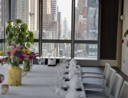 A table set for 14 with white tablecloth and colorful flowers in soft focus, in the background you see the Manhattan cityscape through floor-to-ceiling windows