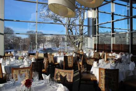 Floor-to-ceiling windows show winter trees and a bright blue sky, round tables and high-backed chairs seat up to 64