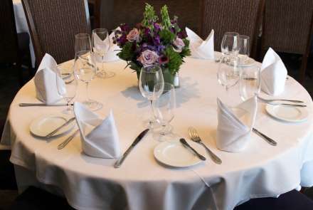 Closeup of a round table set for six, with white tablecloth, cloth napkins, and wineglasses. The centerpiece is an arrangement of lavender roses, purple foxgloves, purple hydrangea, and greenery.