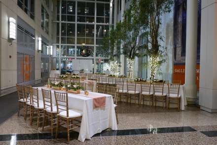 Long tables with white tablecloths and gold chairs, white floral arrangements, potted trees wrapped with white lights, and a stories-high window create the perfect setting for a holiday event