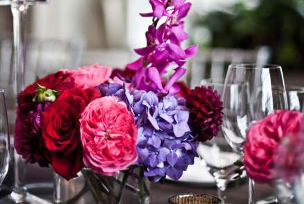 Brightly colored purple, pink, and red flowers are arranged in a glass vase, surrounded by wine glasses and votive candles. A large window is in soft focus in the background.