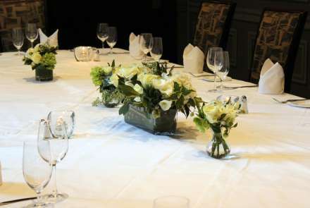 Closeup of white flowers in low vases on a white tablecloth. The table is set with glasses, silverware, and white napkins